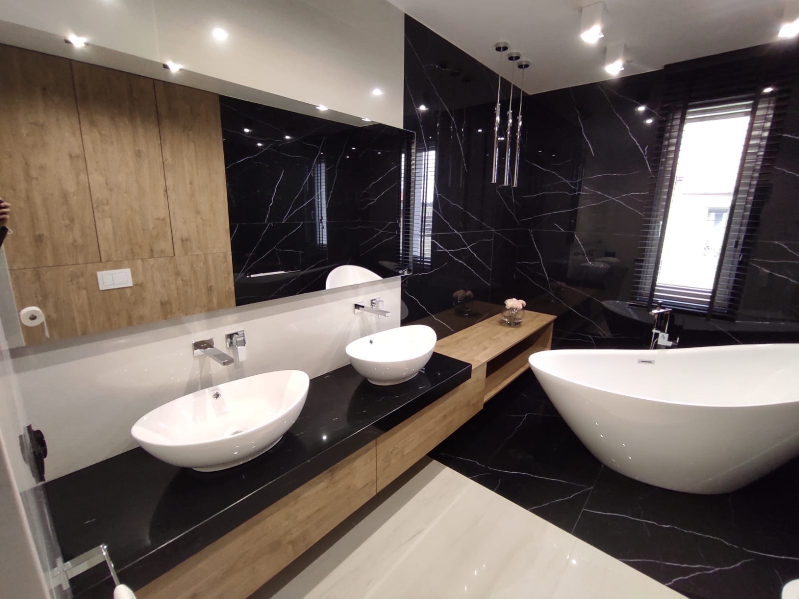 Elegancka łazienka z czarnymi płytkami imitującymi marmur, owalną wanną wolnostojącą i dwiema umywalkami nablatowymi o asymetrycznym kształcie, drewniane szafki pod umywalkami i dekoracyjne...