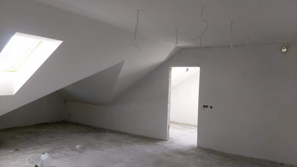 Poddasze po szpachlowaniu i malowaniu na biało, widoczne kable zwisające z sufitu, okno dachowe i surowy beton na podłodze.