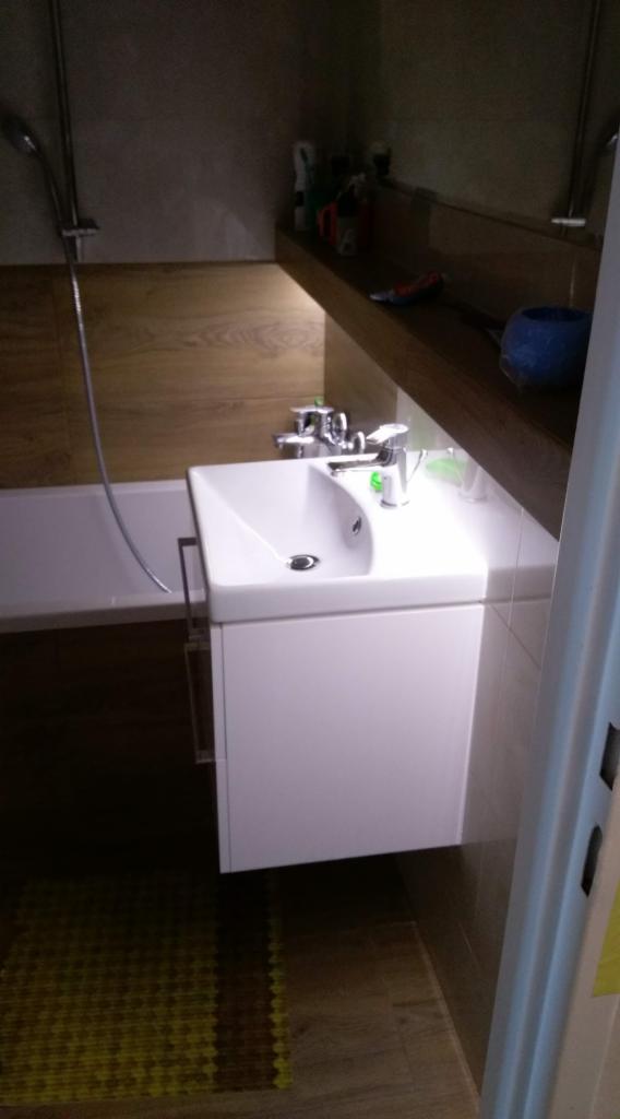 Biała umywalka z szafką w łazience z drewnianymi panelami i półką, widoczna bateria, fragment wanny i mata podłogowa.