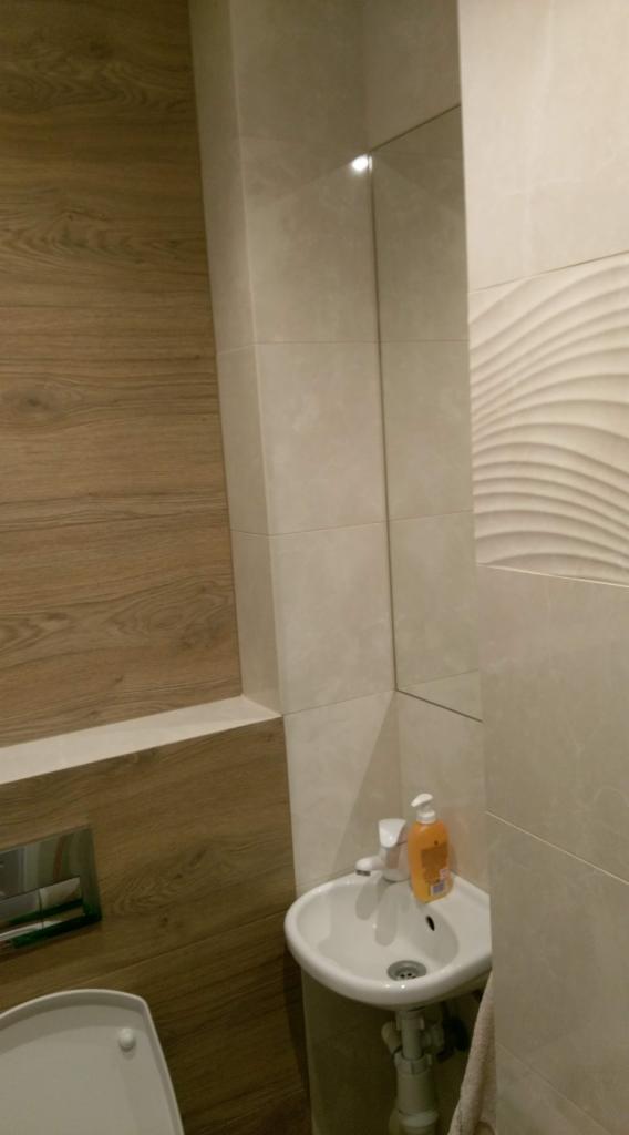 Wykończona toaleta z drewnopodobną okładziną ściany, umywalką narożną z dozownikiem mydła i płytkami ceramicznymi w odcieniach beżu, w tym jedna ściana z płytkami o falistej strukturze.