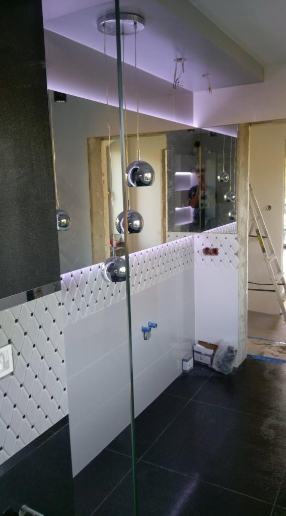 Wykończona łazienka z czarno-białymi płytkami na ścianach, czarną podłogą i trzema chromowanymi lampami wiszącymi, widoczne elementy instalacji i drabina w tle.