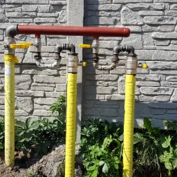 Trzy pionowe rury gazowe w żółtej izolacji, podłączone do poziomego kolektora, na tle szarego muru z kamienia łupanego i zieleni.
