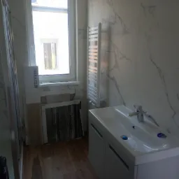 Łazienka w trakcie remontu: kabina prysznicowa, biała umywalka z szafką, grzejnik drabinkowy, marmurowe płytki na ścianach i opakowania płytek Marazzi przy oknie.