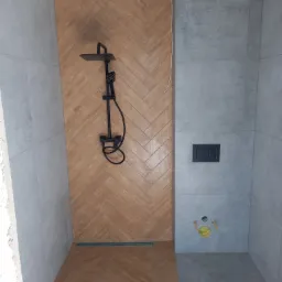 Nowoczesna kabina prysznicowa z drewnopodobnymi płytkami ułożonymi w jodełkę na ścianie, czarna armatura i szare płytki na pozostałych ścianach, widoczny odpływ liniowy.