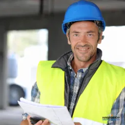 Uśmiechnięty mężczyzna w niebieskim kasku i żółtej kamizelce odblaskowej trzymający plany na budowie.