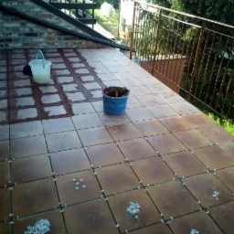 Balkon w trakcie układania płytek ceramicznych w kolorze terakoty, widoczne krzyżyki dystansowe i wiaderka z zaprawą, balustrada w tle.