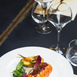 Elegancko podane danie: roladka z farszem, pieczone warzywa (ziemniak, marchew, cukinia), sałata, na białym talerzu, w tle kieliszki do wina i wzorzysty obrus.