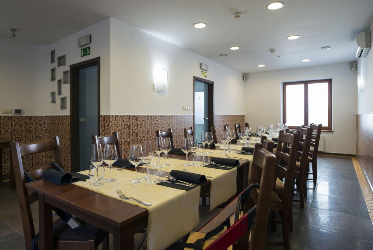 Sala restauracyjna przygotowana do przyjęcia gości, z długim stołem nakrytym jasnym obrusem, ciemnymi serwetkami, kieliszkami i sztućcami, w tle wzorzyste panele ścienne i okno.