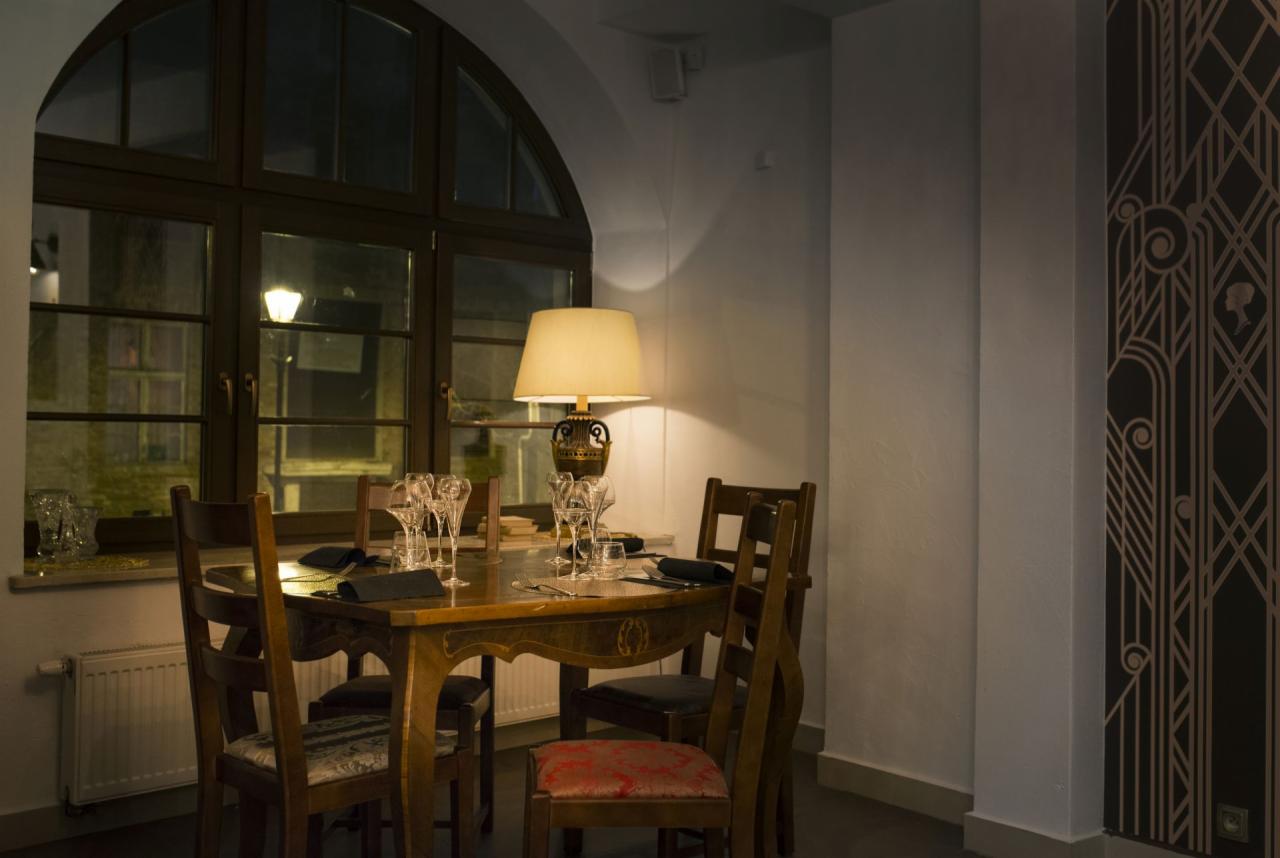 Elegancko nakryty stół w restauracji z ciemnymi drewnianymi krzesłami, kryształowymi kieliszkami i czarnymi serwetkami, oświetlony lampą ze stożkowym abażurem, z widokiem na nocne miasto przez...