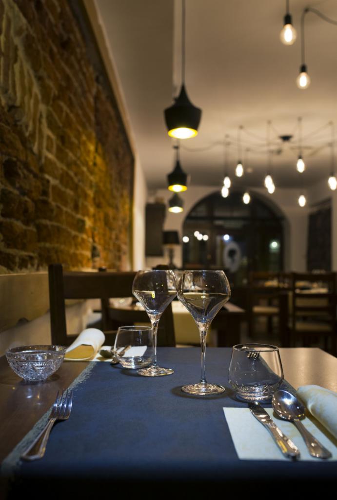 Elegancko nakryty stół w restauracji z dwoma kieliszkami do wina, szklankami, sztućcami i serwetkami na tle ceglanej ściany i dekoracyjnych lamp.