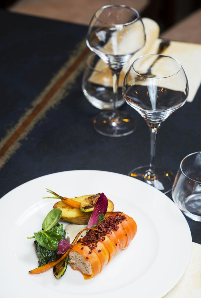 Elegancko podane danie: roladka z farszem, pieczone warzywa (ziemniak, marchew, cukinia), sałata, na białym talerzu, w tle kieliszki do wina i wzorzysty obrus.