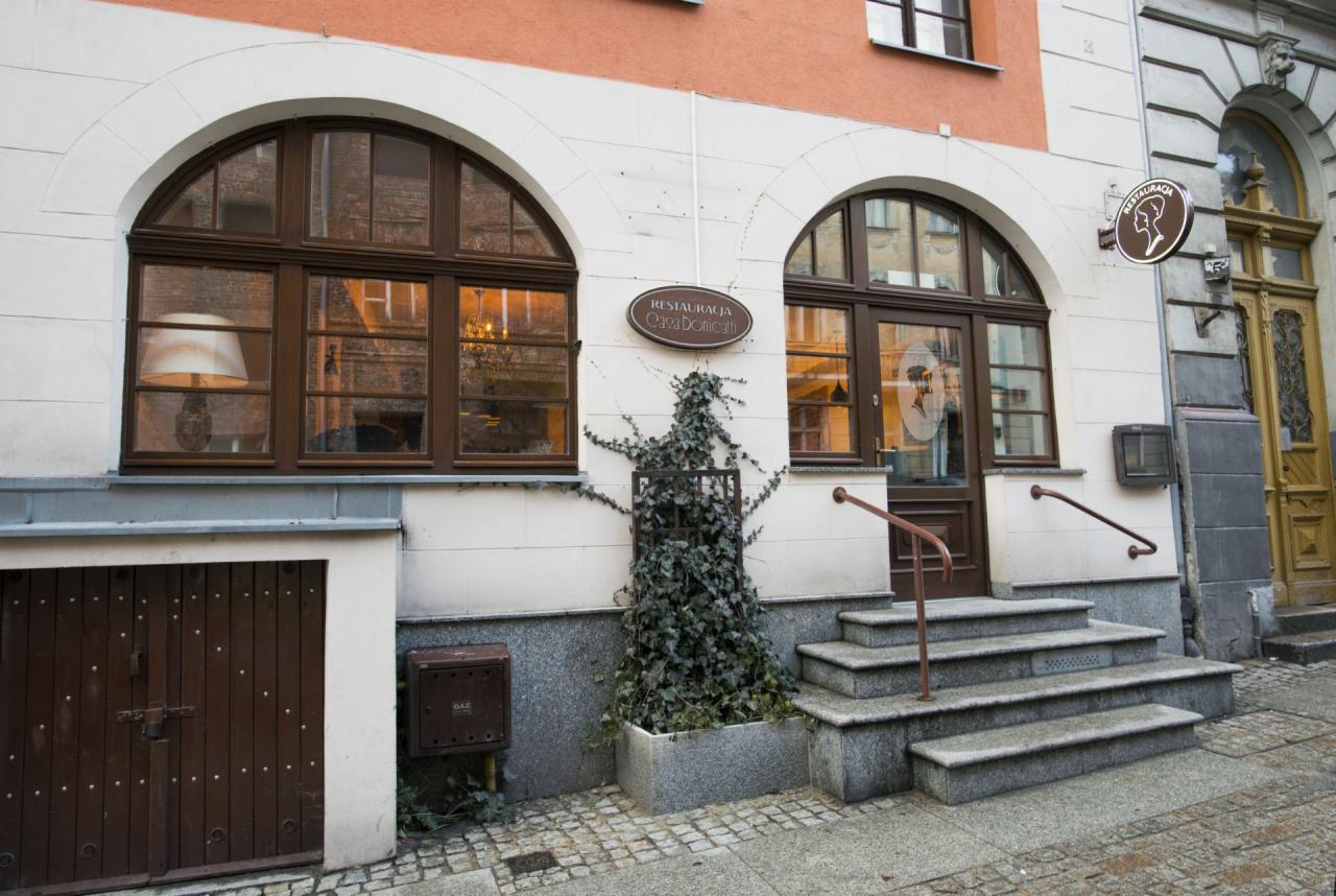 Wejście do restauracji z marmurowymi schodami, przeszklonymi drzwiami z brązowymi ramami, oknami łukowymi i tabliczką z nazwą 'Restauracja Casa Domnicati' oraz donicą z bluszczem.