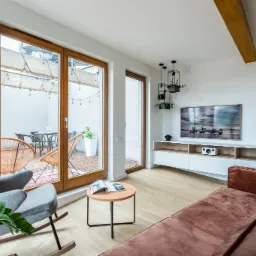 Jasne wnętrze salonu z drewnianą podłogą, brązową sofą i szarym fotelem bujanym, z widokiem na taras z drewnianą podłogą i meblami ogrodowymi przez przeszklone drzwi.