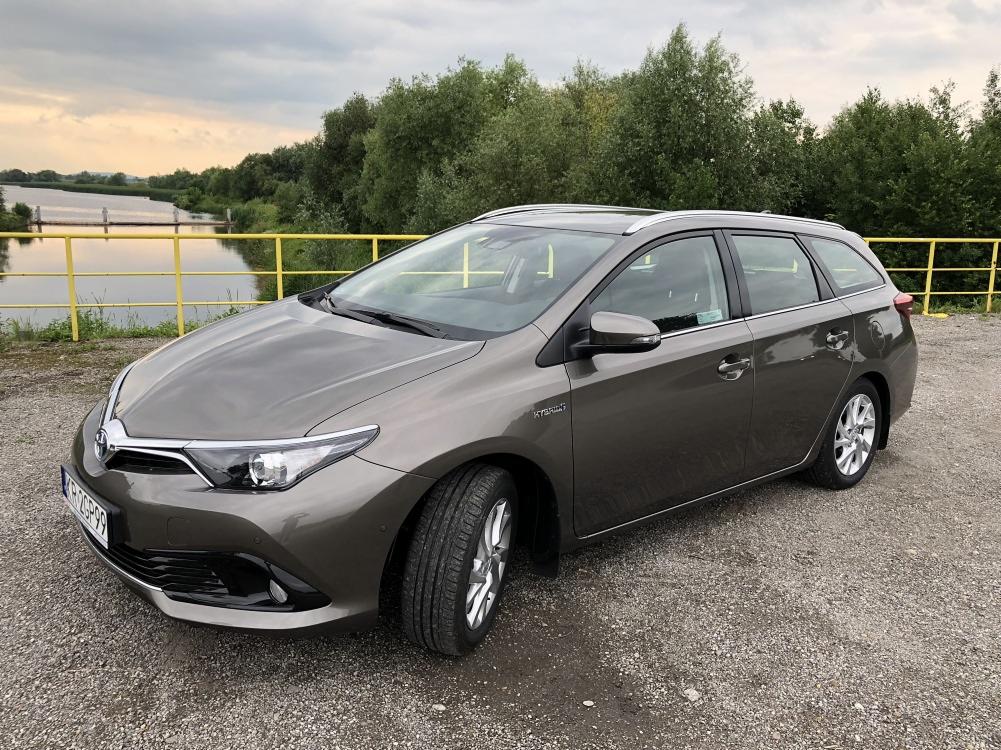 Szary samochód kombi Toyota Auris Hybrid zaparkowany na żwirowym podłożu z widokiem na rzekę i drzewa w tle.