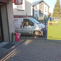 Żółty baner reklamowy z napisem 'Finansowanie Twoich potrzeb, Produkty bankowe i pozabankowe' przed budynkiem, częściowo zasłonięty przez metalowe ogrodzenie, za którym stoi biały samochód dostawczy.