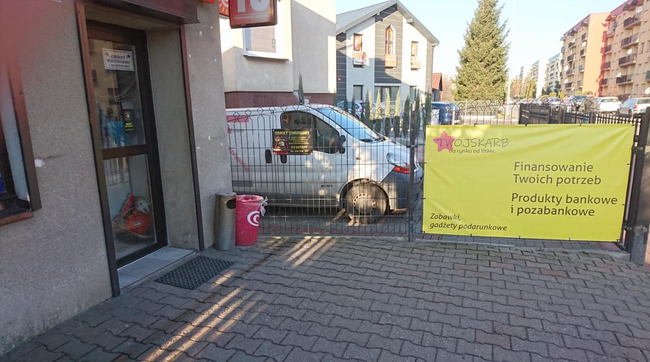Żółty baner reklamowy z napisem 'Finansowanie Twoich potrzeb, Produkty bankowe i pozabankowe' przed budynkiem, częściowo zasłonięty przez metalowe ogrodzenie, za którym stoi biały samochód dostawczy.