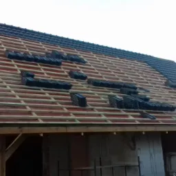 Montaż czarnej dachówki na drewnianej konstrukcji dachu domu w trakcie budowy, widoczne ułożone stosy dachówek i fragmenty gotowej połaci.