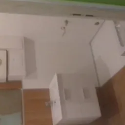 Biała łazienka z brązowymi płytkami podłogowymi, widoczny bojler, umywalka z szafką i fragment wanny. Na ścianie widoczne ślady po remoncie.