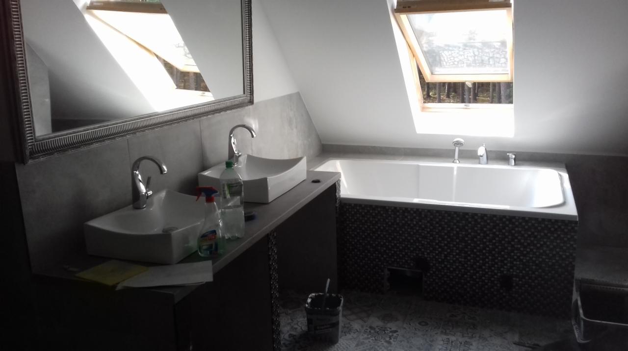 Dwie białe umywalki o nowoczesnym designie z chromowanymi kranami w łazience na poddaszu, widoczne środki czystości i okno dachowe nad wanną z mozaiką.