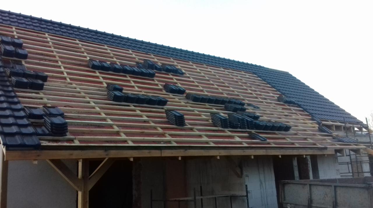 Montaż czarnej dachówki na drewnianej konstrukcji dachu domu w trakcie budowy, widoczne ułożone stosy dachówek i fragmenty gotowej połaci.