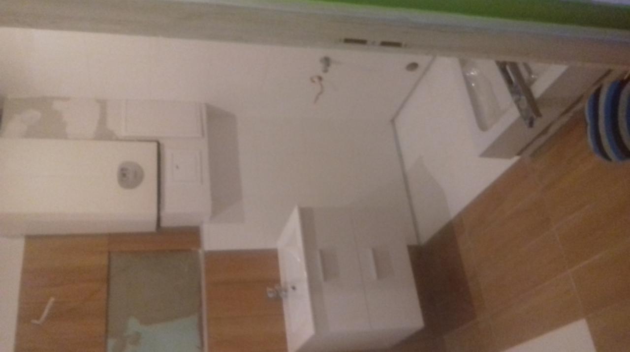 Biała łazienka z brązowymi płytkami podłogowymi, widoczny bojler, umywalka z szafką i fragment wanny. Na ścianie widoczne ślady po remoncie.