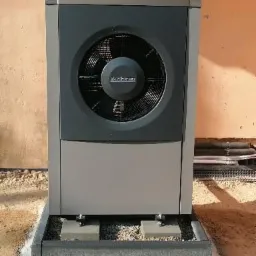 Szara pompa ciepła marki Buderus zamontowana na betonowej podstawie, widok z przodu na tle ściany budynku.