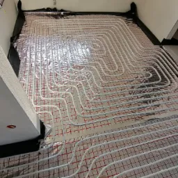 Białe rury ogrzewania podłogowego ułożone na srebrnej folii z czerwoną siatką, widoczne czarne pasy izolacji przy ścianach w surowym wnętrzu.