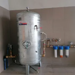 Srebrny, pionowy zbiornik hydroforowy Wimest ZCS/300/6 z manometrem, podłączony do instalacji z trzema filtrami wody o niebieskich obudowach, zamontowany na brudnej, kafelkowej podłodze...