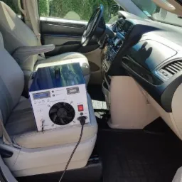 Urządzenie do ozonowania wnętrza samochodu umieszczone na fotelu pasażera, widoczny kabel zasilający i panel sterowania.