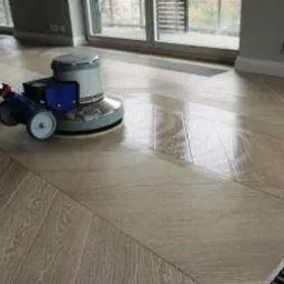 Automat szorujący poleruje parkiet ułożony we wzór jodełki, widoczne refleksy światła na mokrej powierzchni, w tle okno i fragment ściany z listwą przypodłogową.