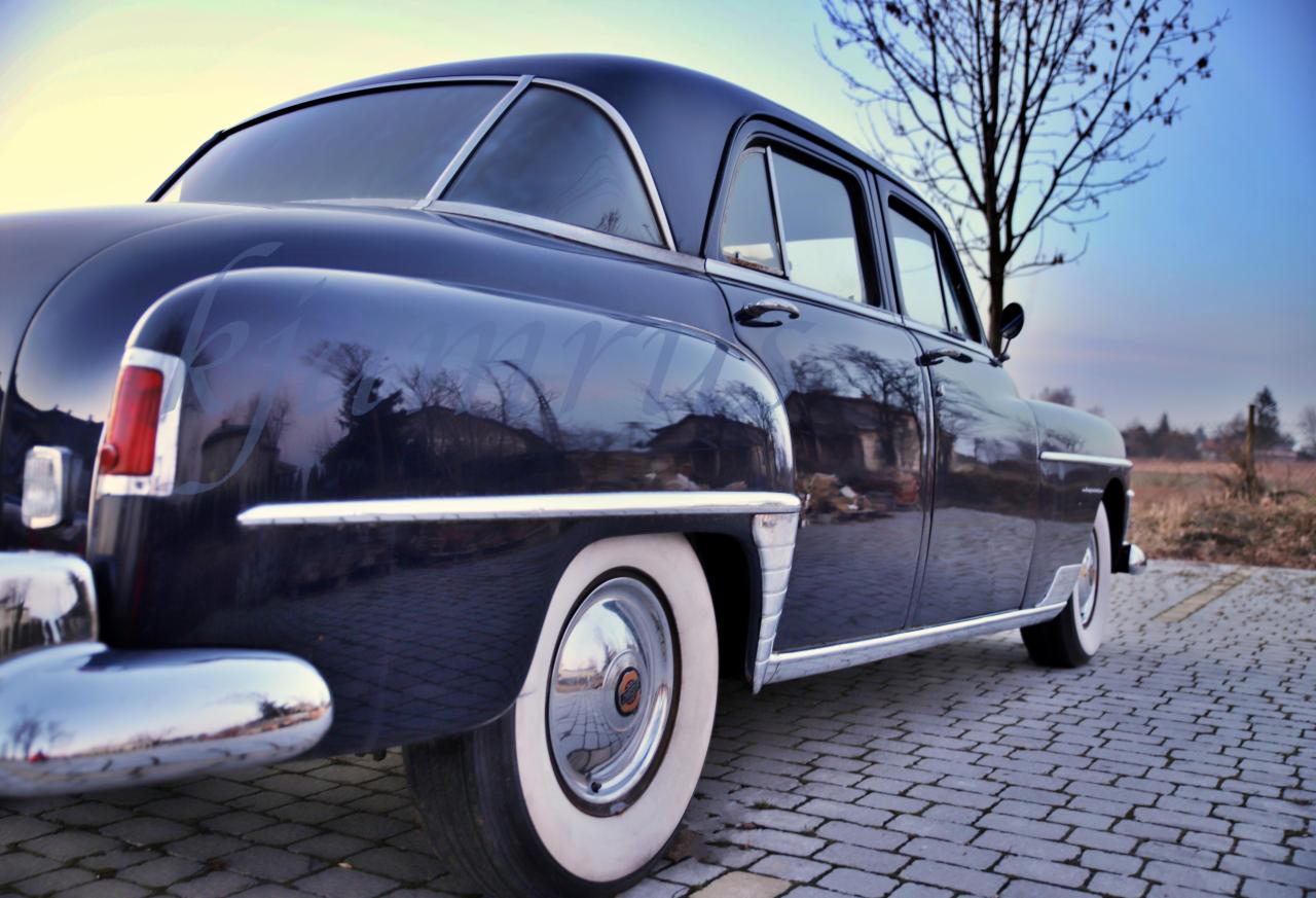 Czarny, zabytkowy samochód z białymi oponami, zaparkowany na brukowanej drodze, z odbiciem budynków w karoserii.