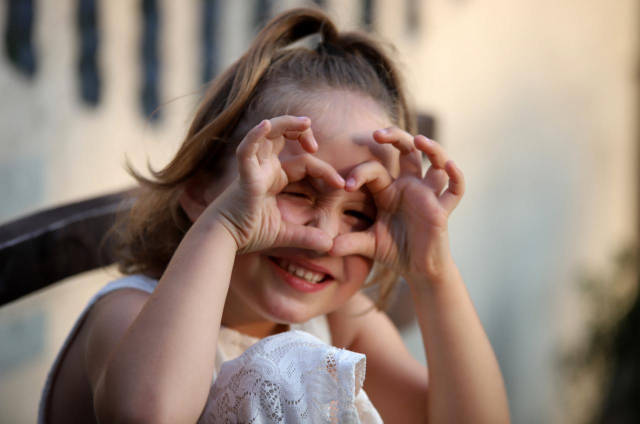 Uśmiechnięta dziewczynka w białej sukience, siedząc, formuje dłońmi kształt serca na tle rozmytego otoczenia.