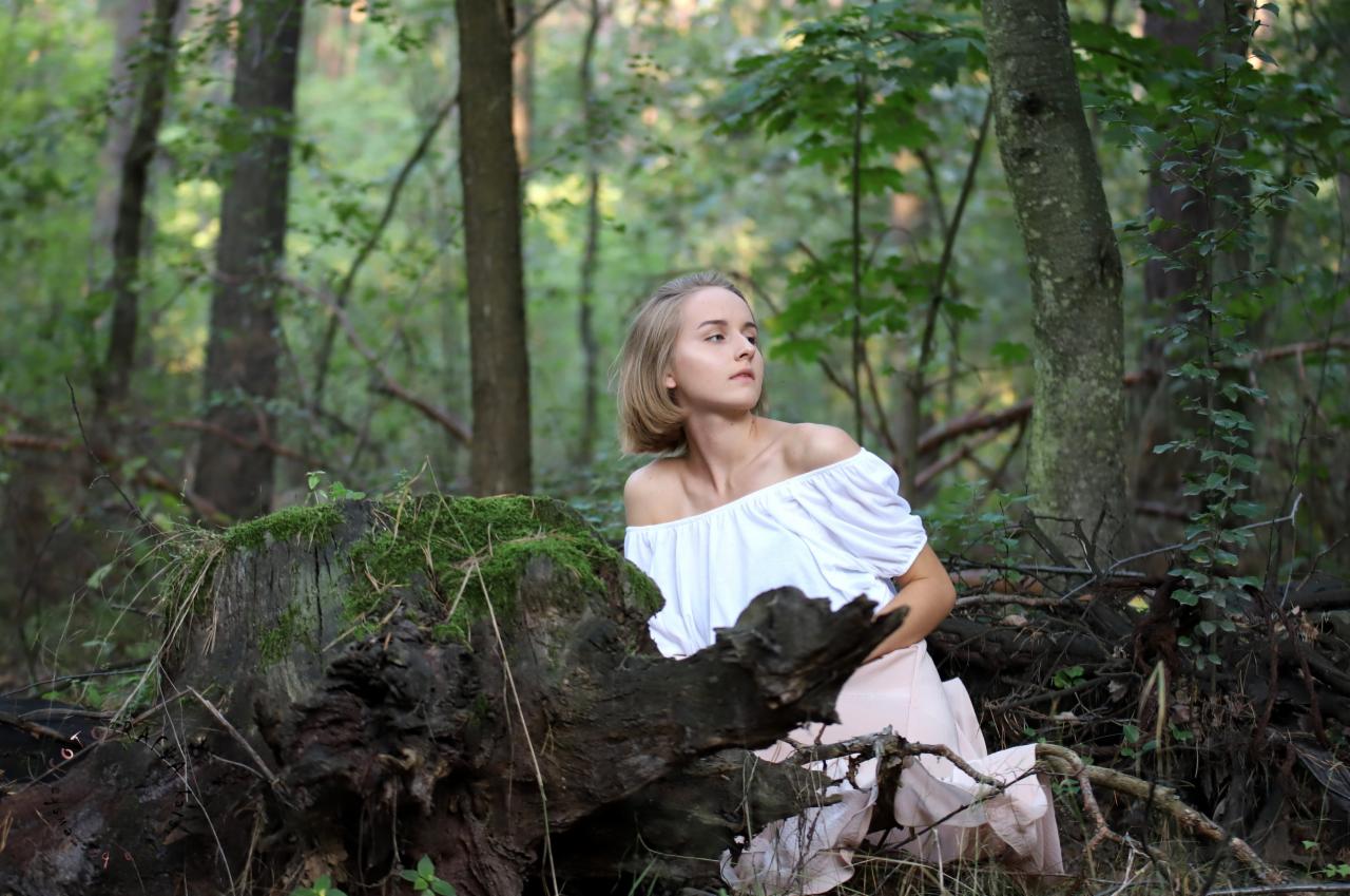 Młoda kobieta w białej bluzce z odkrytymi ramionami i różowej spódnicy siedzi w lesie opierając się o zwalony pień drzewa porośnięty mchem, spoglądając w dal.