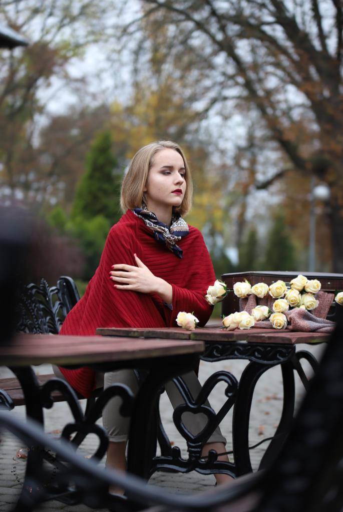 Portret kobiety w czerwonym ponczo siedzącej przy stoliku z białymi różami i walizką w jesiennym parku.