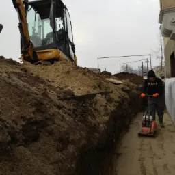 Wykop szerokoprzestrzenny, ubijarka spalinowa w akcji, mini koparka na skraju wykopu, przygotowanie terenu pod fundamenty, pochmurny dzień.