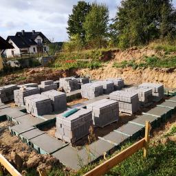 WAKO - dom to podstawa - Fundamenty pod dom z betonu, z przygotowanymi miejscami na ściany z bloczków betonowych, widok z góry na teren budowy z zielenią i budynkami w tle.