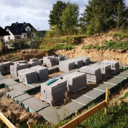 Fundamenty pod dom z betonu, z przygotowanymi miejscami na ściany z bloczków betonowych, widok z góry na teren budowy z zielenią i budynkami w tle.