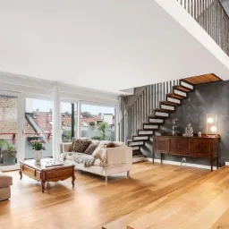 Jasne, przestronne wnętrze z drewnianą podłogą, schodami z metalową balustradą, sofą z poduszkami, stolikiem kawowym i widokiem na balkon z roślinami.