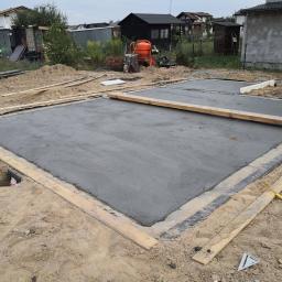 WAKO - dom to podstawa - Wylana betonowa płyta fundamentowa pod budynek, otoczona drewnianym szalunkiem i folią izolacyjną, z widoczną pomarańczową betoniarką w tle.