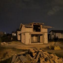 WAKO - dom to podstawa - Niedokończony dwukondygnacyjny dom z pustaków, sfotografowany nocą, z widocznym stosem desek na pierwszym planie i innymi budynkami w tle.