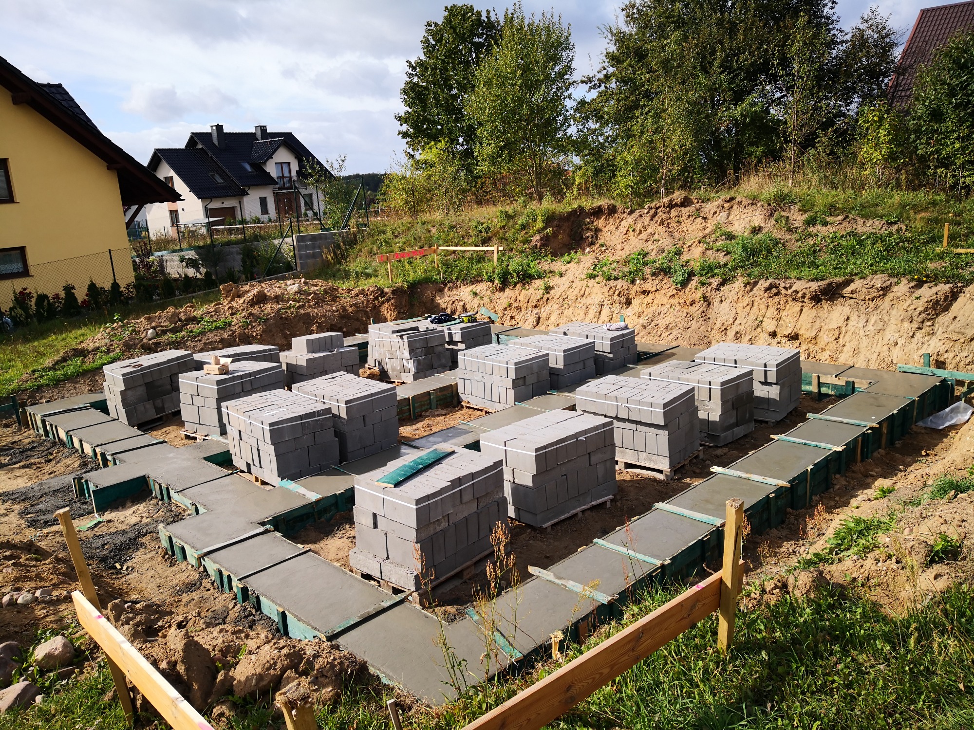 Fundamenty pod dom z betonu, z przygotowanymi miejscami na ściany z bloczków betonowych, widok z góry na teren budowy z zielenią i budynkami w tle.