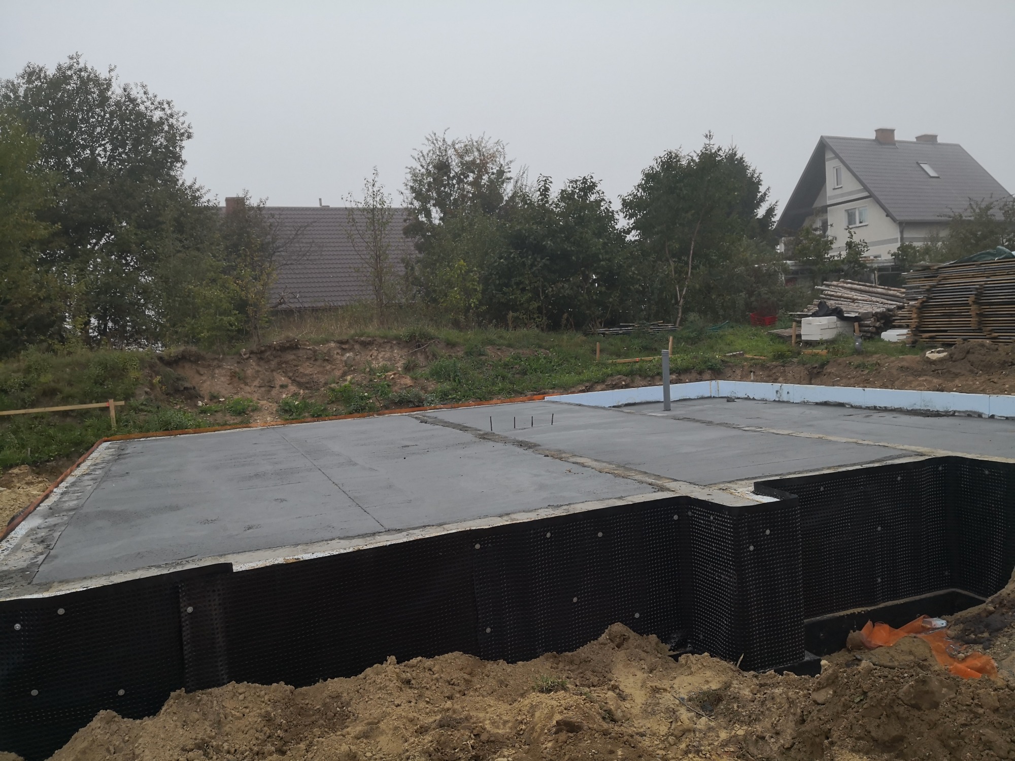 Wylana płyta fundamentowa z izolacją pionową ścian fundamentowych, widoczne wystające pręty zbrojeniowe, w tle dom w budowie i drzewa.