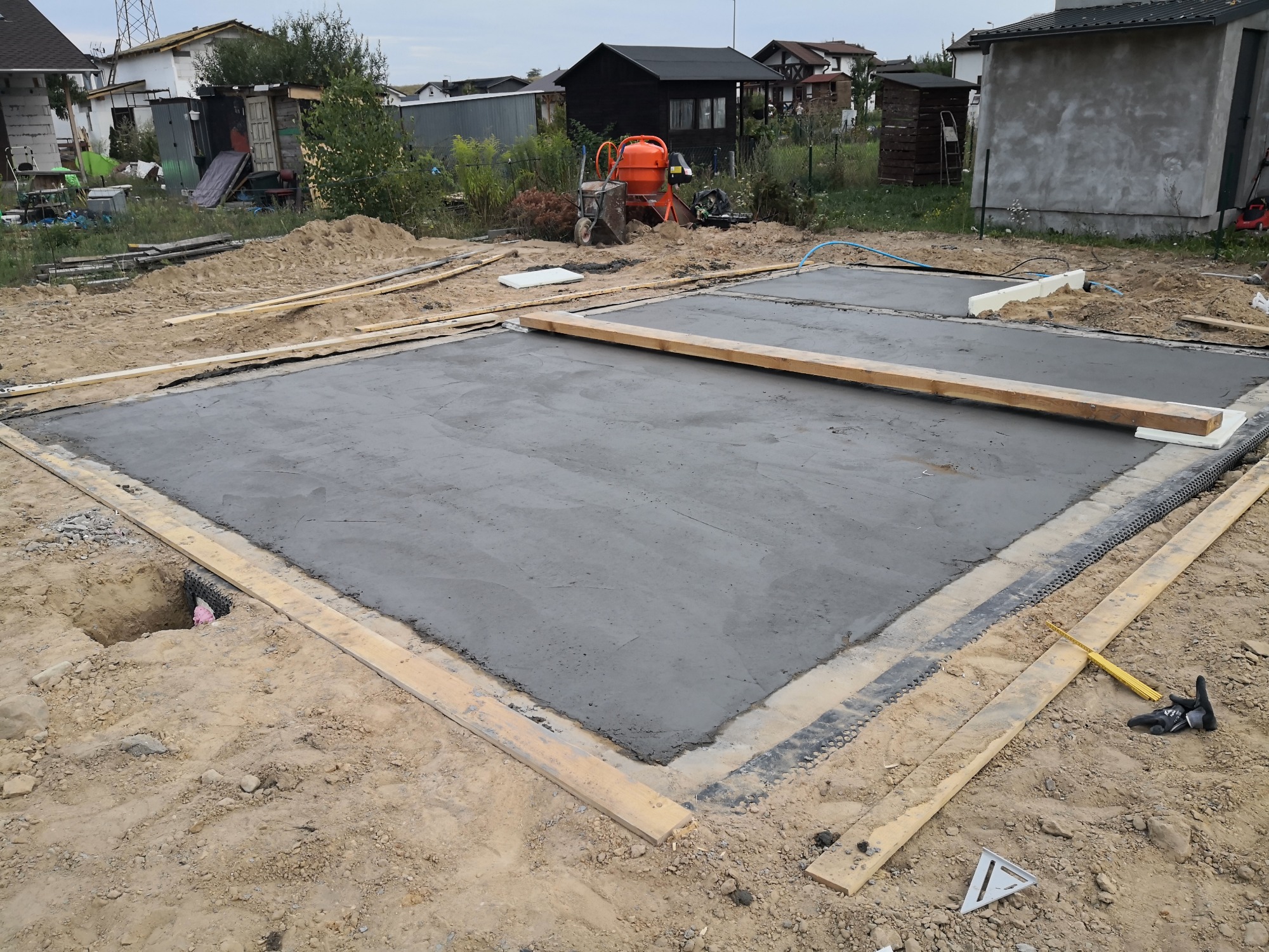 Wylana betonowa płyta fundamentowa pod budynek, otoczona drewnianym szalunkiem i folią izolacyjną, z widoczną pomarańczową betoniarką w tle.