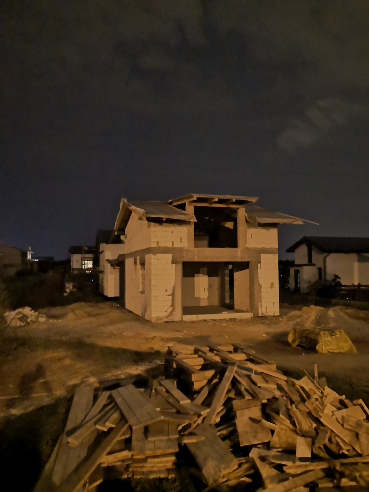 Niedokończony dwukondygnacyjny dom z pustaków, sfotografowany nocą, z widocznym stosem desek na pierwszym planie i innymi budynkami w tle.