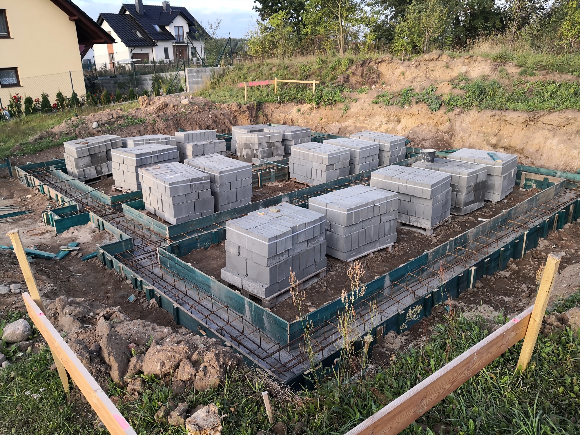 Przygotowany fundament pod dom z szalunkiem i zbrojeniem, na którym ułożone są palety z bloczkami betonowymi.