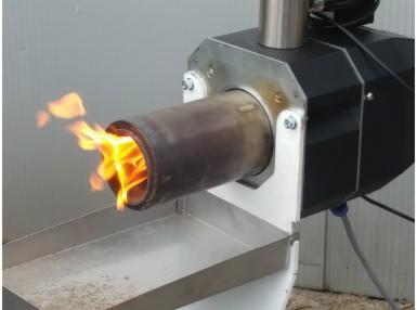 Płomienie wydobywające się z metalowego palnika pieca na paliwo stałe z widocznym popielnikiem, na tle szarej ściany.