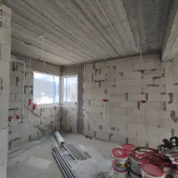 Surowy pokój w trakcie budowy z widocznymi przewodami elektrycznymi w peszlach na ścianach z betonu komórkowego, puszki elektryczne zamontowane, okno z widokiem na zewnątrz, na podłodze materiały...