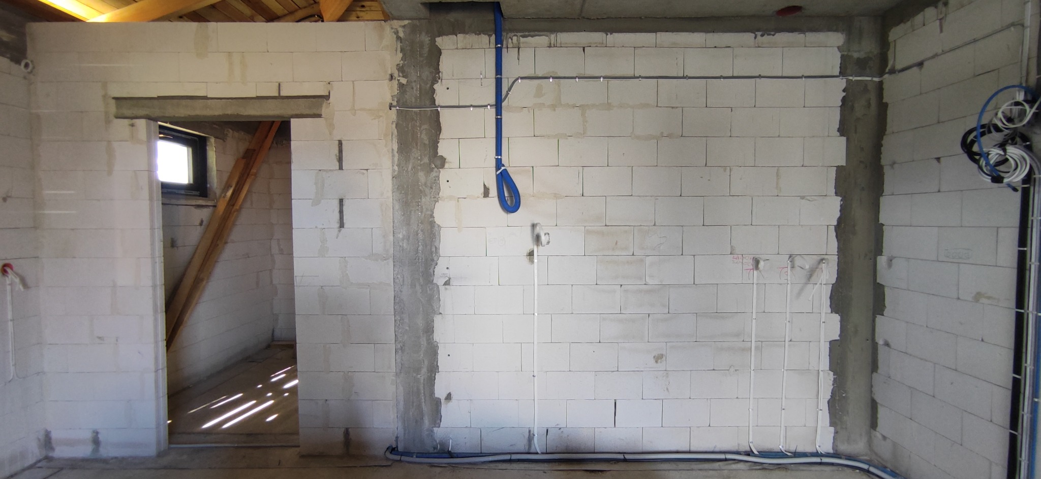 Surowy stan wnętrza budynku w trakcie prac instalacyjnych: widoczne przewody elektryczne prowadzone po ścianach z betonu komórkowego oraz niebieska rura karbowana. W tle surowe drewniane elementy...