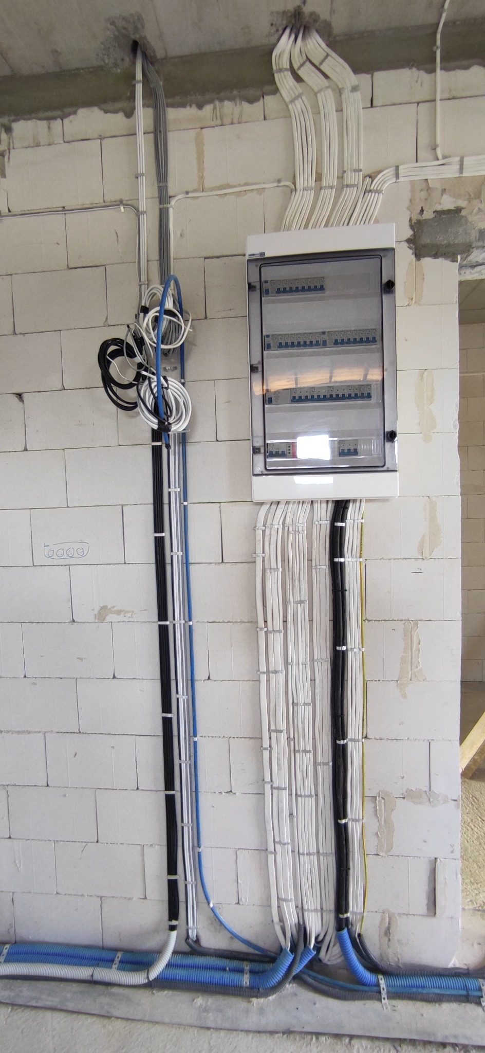 Rozdzielnica elektryczna z zabezpieczeniami, do której podłączone są liczne przewody w kolorach białym, czarnym i niebieskim, przymocowane do ściany z białych bloczków.