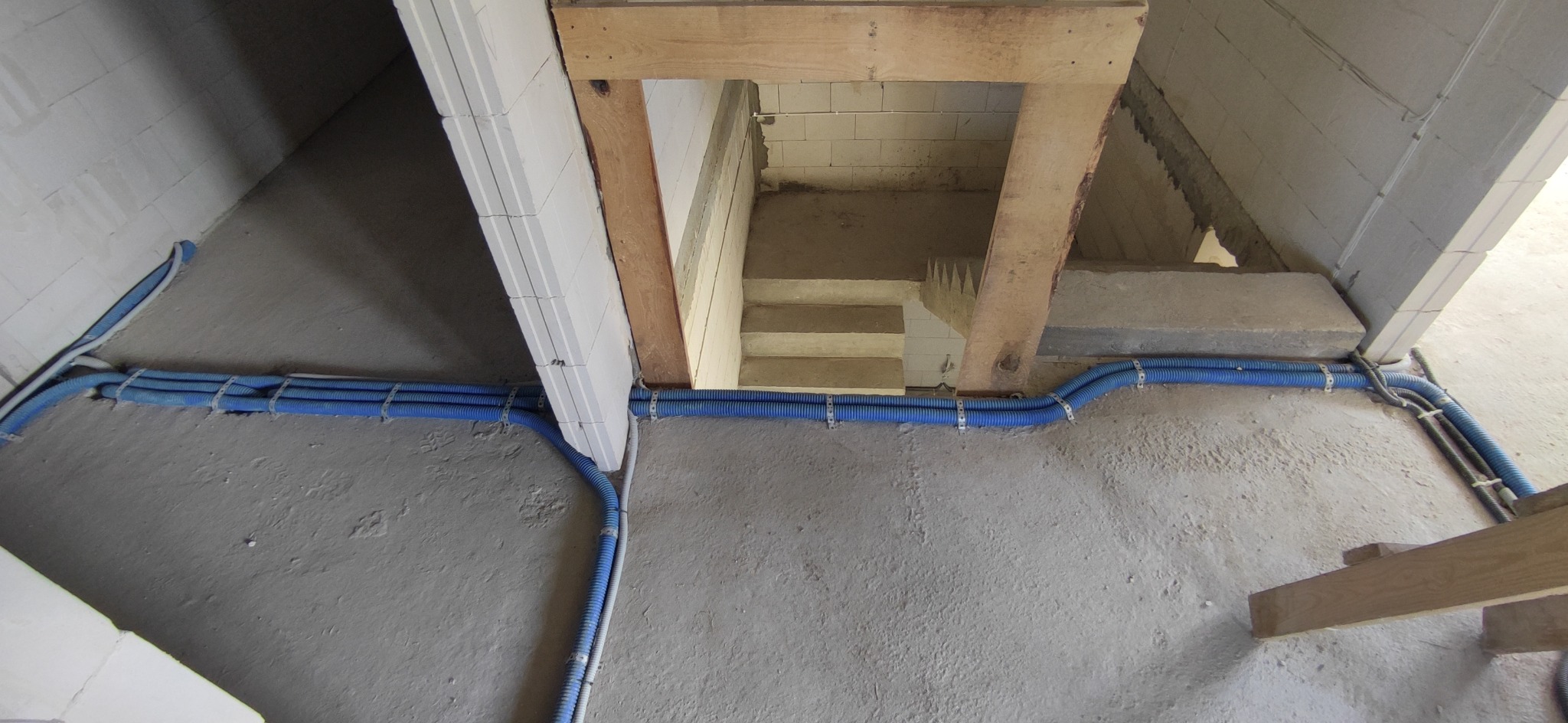 Niebieskie rury karbowane prowadzone wzdłuż ściany z betonu komórkowego w nowo budowanym domu, widoczne drewniane elementy konstrukcyjne oraz betonowe schody w tle.